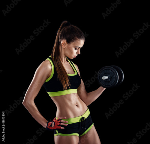 Young athletic woman in sportswear with dumbbells in studio against black background. Ideal female sports figure. Fitness girl with perfect sculpted muscular and tight body.