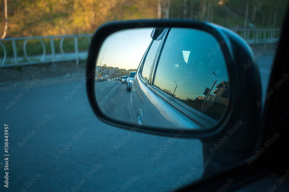 Row of cars on road is visible in side-view mirror of car in summer evening.