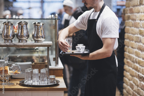Waiter wearing apron and holding tray with cup of coffee and a glass.