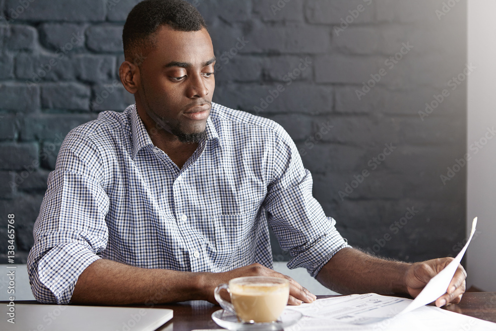 Candid indoor shot of attractive dark-skinned businessman in checkered shirt holding document and reading information with concentrated serious look, reviewing his accounts during lunch at cafe Candid indoor shot of attractive dark-skinned businessman in checkered shirt holding document and reading information with concentrated serious look, reviewing his accounts during lunch at cafe
