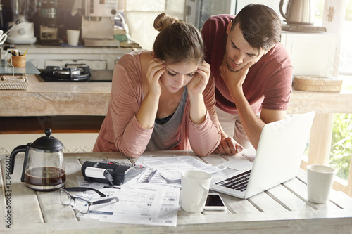 Stressed young Caucasian couple facing financial problems, not able to pay off their loan because of many debts. Woman with headache and worried man feeling stress, reading notification from bank