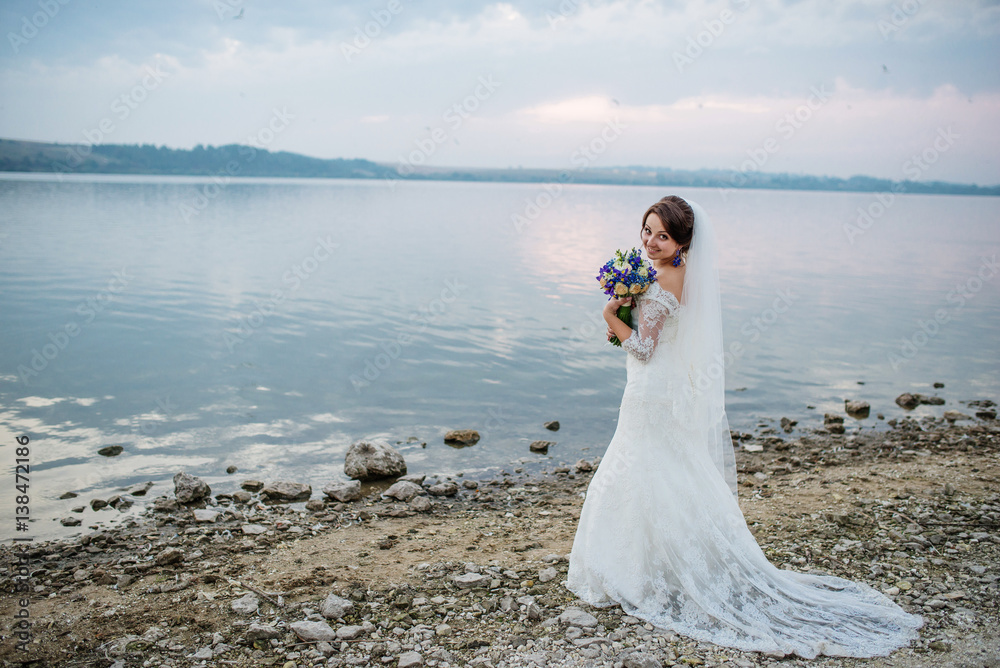 Brunette bride with violet wedding bouquet near beach of river at sunset. Brunette bride with violet wedding bouquet near beach of river at sunset.