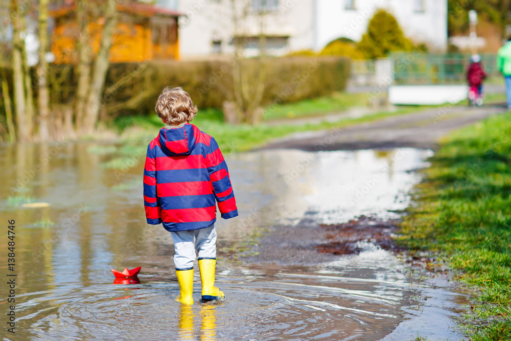 Little kid boy playing with paper ship by puddle Little kid boy playing with paper ship by puddle
