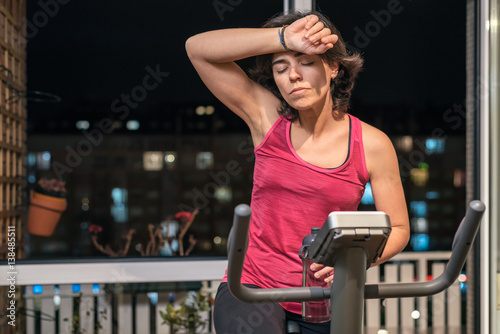 Fotografia Healthy fit sporty woman exhausted and dizzy rising forearm to forehead at home on exercise static bike after daily female workout routine