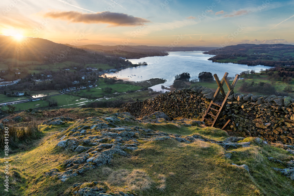 Fototapeta premium Sun rising above the horizon illuminating the landscape with stone wall and stile in foreground.