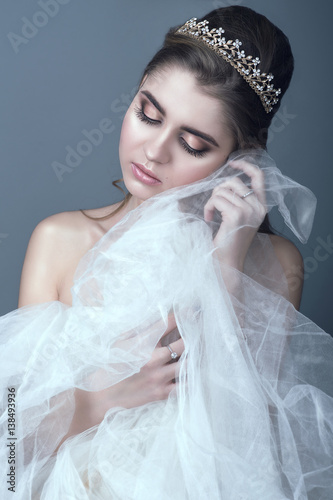 Portrait of young beautiful bride in diadem with naked shoulders touching fluffy skirt of her wedding dress with her cheek, her eyes closed. Isolated on grey background. Studio shot
