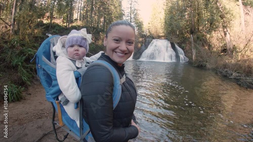Mother Smiling and Waving at Camera with Baby Daughter in Hiking Backpack at Forest Waterfall