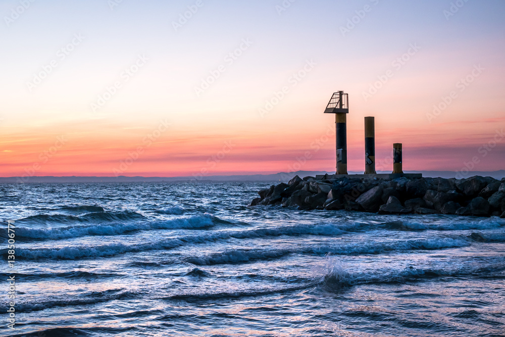 paysage au coucher de soleil sur la mer avec une digue et trois structures métalliques au bout dont une guérite
