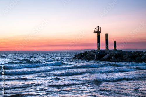 paysage au coucher de soleil sur la mer avec une digue et trois structures métalliques au bout dont une guérite