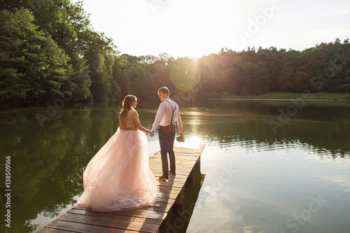 Wedding couple walking on bridge near lake on sunset at wedding day. Bride and groom in love
