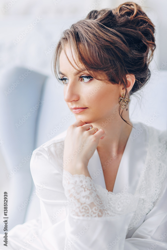 Close up portrait of young beautiful bride in elegant white robe sitting on a chair in a bright interior with a fireplace on the background Close up portrait of young beautiful bride in elegant white robe sitting on a chair in a bright interior with a fireplace on the background