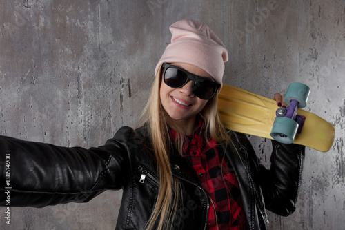 Blond teenager girl in black sunglasses and pink hat with yellow skateboard make selfie. In front of concrete wall.