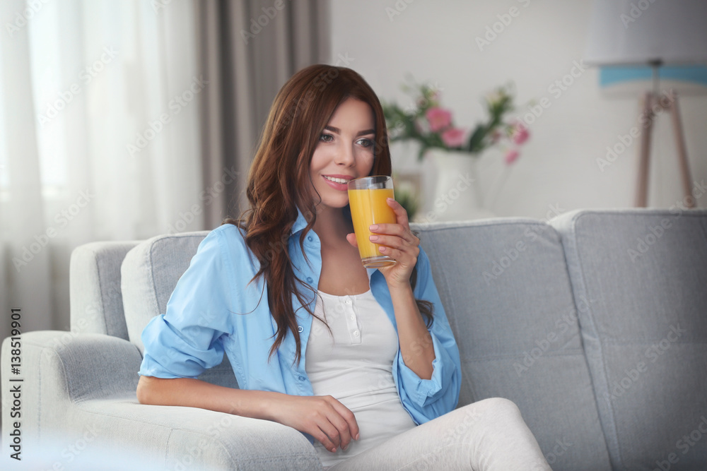 Beautiful young woman with glass of orange juice sitting on sofa at home
