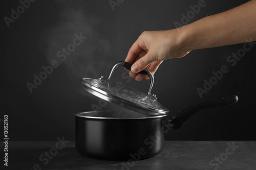 Human hand removing cover from saucepan with hot liquid, on dark background