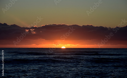 Beautiful sunrise cloudscape over ocean background. Sun rays beaming through picturesque clouds above sea. Blue sky with clouds, sea and sun on the horizon. Beautiful sunrise over the quiet calm sea.