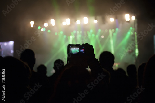 Close up on hands taking photos with phone on a concert, colorful stage in the background.