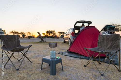 Woman cooking with gas stove in camping site at dusk. Gas burner, pot and smoke from boiling water, tent in the background. Adventures in african national parks.