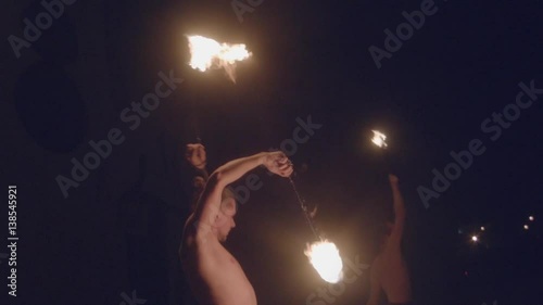 Fire show street circus performance close up slow motion. Two male fire performers spinning burning fire poi at black background in night during carnival photo