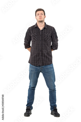 Serious suspicious man with hands behind back looking at camera. Full body length portrait isolated over white background.