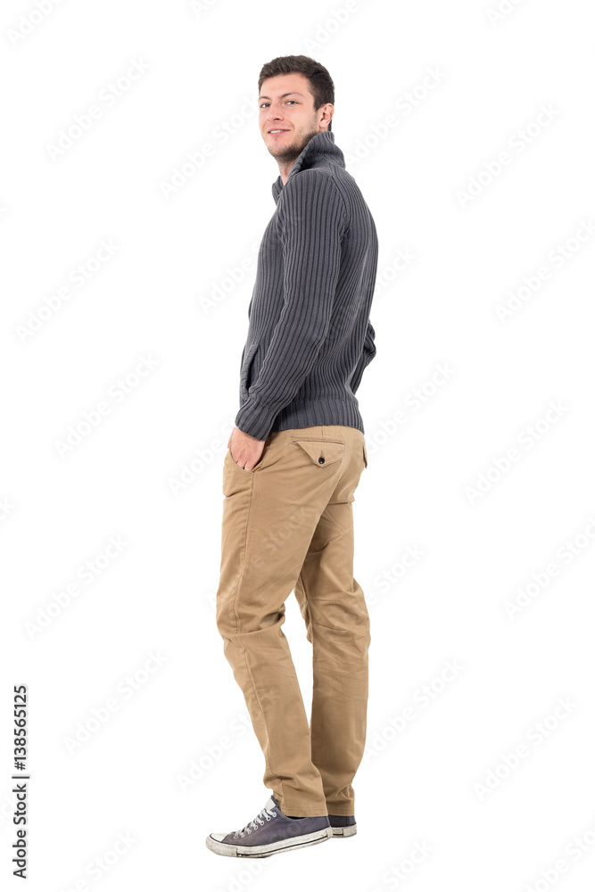 Rear view of young man in jumper with hands in pockets smiling at camera. Full body length portrait isolated over white background. Rear view of young man in jumper with hands in pockets smiling at camera. Full body length portrait isolated over white background.