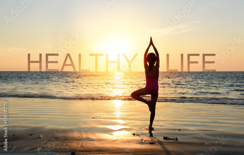  Silhouette of A girl practicing Yoga vrikshasana tree pose on tropical beach with sunset sky background, watching the sunset, standing as a part of the wording concept for healthy life. 