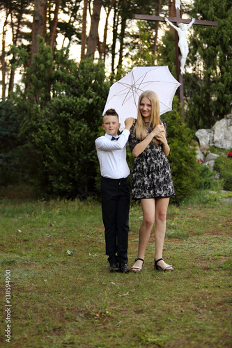 Siostra i brat stoją uśmiechnięci pod parasolem w parku.