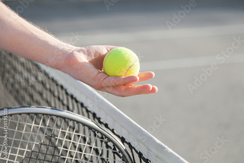 Sportsman is playing tennis on court outdoor. Hand gives yellow tennis ball over net to other player. Athlete holds tennis racket. © Marina April