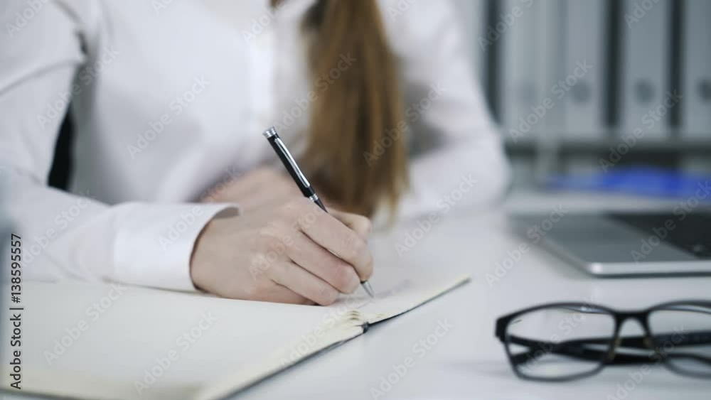 Female hand writing on a notepad with shallow depth of field. Laptop and glasses are on the desk. Locked down real time close up shot. Female hand writing on a notepad with shallow depth of field. Laptop and glasses are on the desk. Locked down real time close up shot.