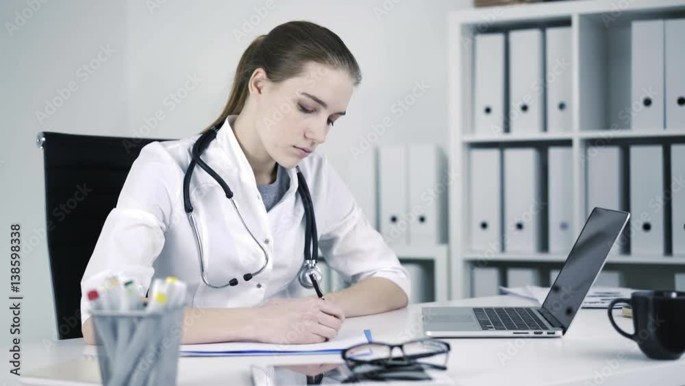 Beautiful general practicer is sitting on the chair at medical office and writing down a prescription and diagnosis details in a report book. Locked down real time medium shot. Beautiful general practicer is sitting on the chair at medical office and writing down a prescription and diagnosis details in a report book. Locked down real time medium shot.