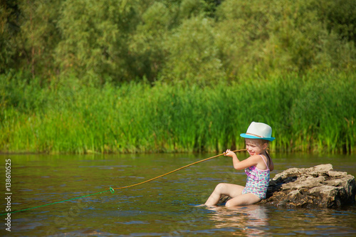 Fototapeta Naklejka Na Ścianę i Meble -  Cute little girl in funny hat pretends to catch fish, child plays fishing on river