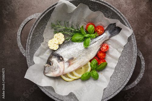 Fresh uncooked dorado or sea bream fish with lemon, herbs, oil, vegetables and spices in a frying pan on a baking paper over black backdrop, top view