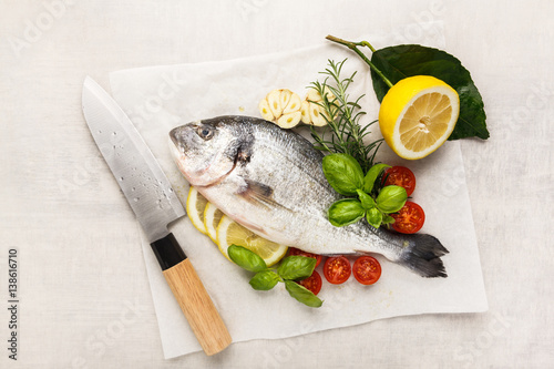 Fresh uncooked dorado or sea bream fish with lemon, herbs, oil, vegetables and spices in a frying pan on a baking paper over white backdrop, top view