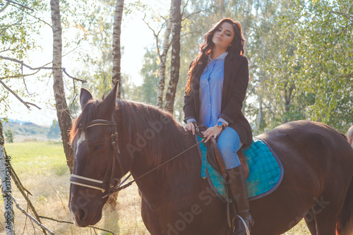 Beautiful sexy brunette wearing jeans, blouse and black jacket riding a horse at sunny summer day