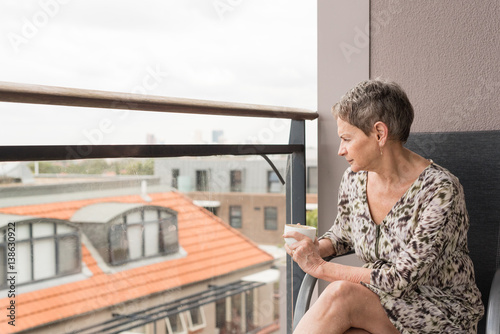 Older woman seated on balcony with coffee cup against urban background (selective focus)