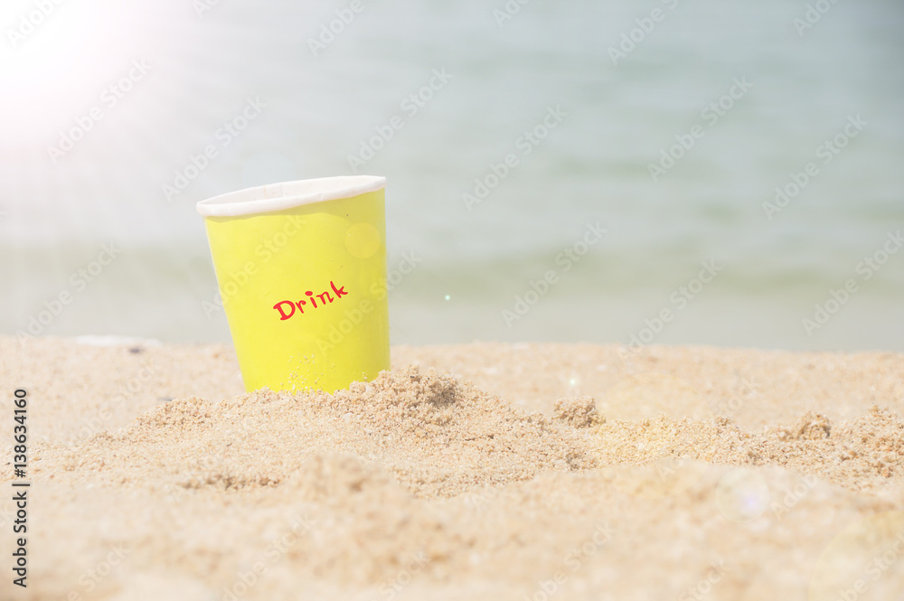 Yellow paper glass beverage on the sand behind beach with sunlight.