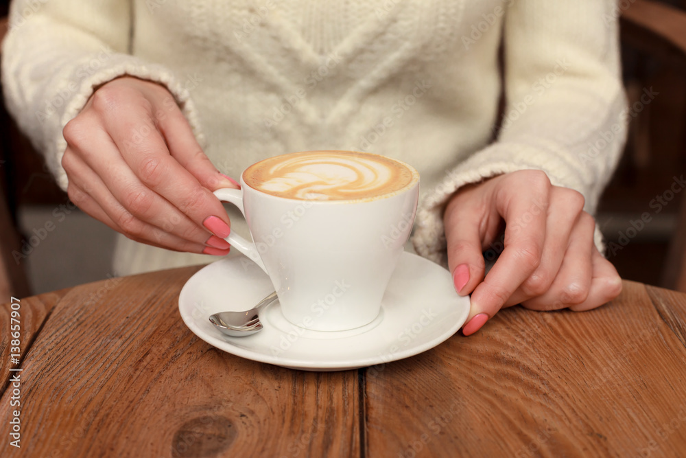 Close-up female hands holding cup with coffee cappuccino with foam with nice pattern. Perfect red gel polish manicure. Wood natural vintage table. Creative color warm post processing Close-up female hands holding cup with coffee cappuccino with foam with nice pattern. Perfect red gel polish manicure. Wood natural vintage table. Creative color warm post processing