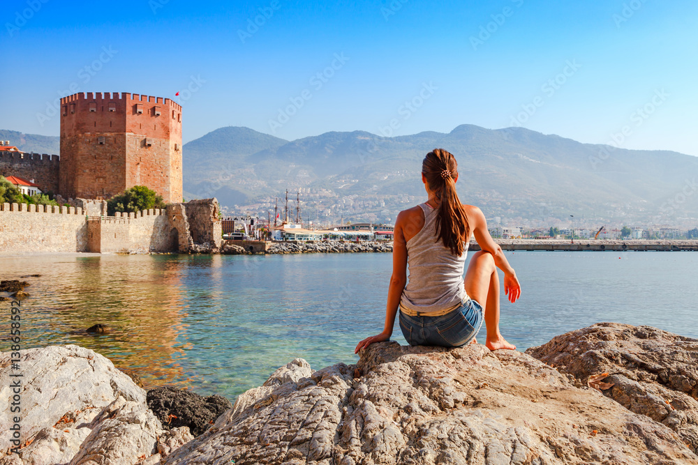 Fototapeta premium Young woman look at Kizil Kule tower in Alanya peninsula, Antalya district, Turkey, Asia. Famous tourist destination with high mountains. Part of ancient old Castle. Summer bright day