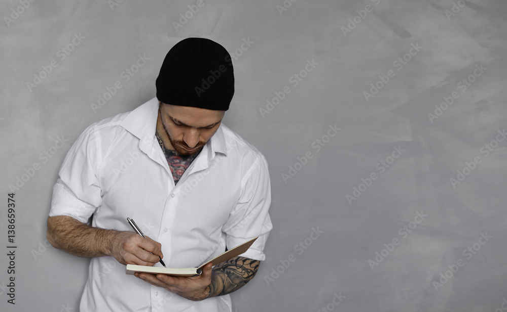 Handsome latin man in white jacket and black hat stay near grey wall and write notes in notepad. Attractive student writing in drawing book. Studio shot with copy space for advertising text.