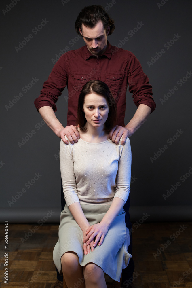 Young couple, woman is sitting on the chair, the man is standing behind,  his hands keeping her shoulders, gray background Stock-Foto | Adobe Stock