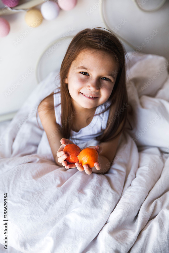 Little beautiful girl with tangerines in hands and in bed. Home entertainment, recreation Little beautiful girl with tangerines in hands and in bed. Home entertainment, recreation