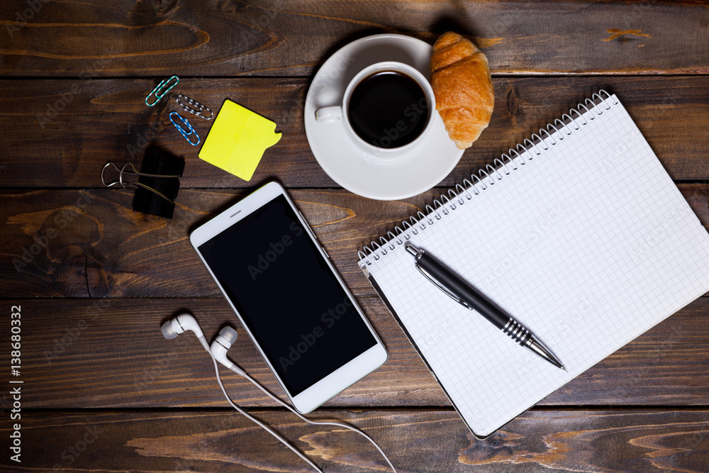 White mobile phone on wooden wooden background with headphones, cup of coffee, croissant and Stationery appliances White mobile phone on wooden wooden background with headphones, cup of coffee, croissant and Stationery appliances