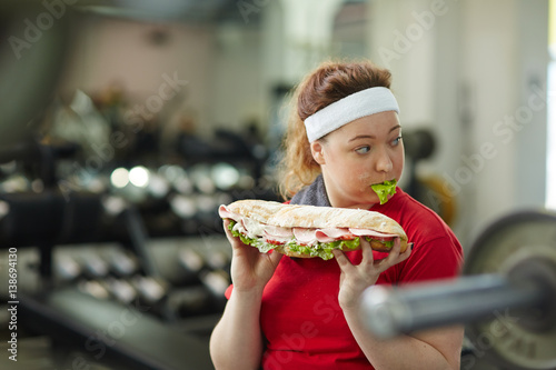 Portrait of young overweight woman eating big greasy fattening sandwich at work out in gym, concept of food obsession