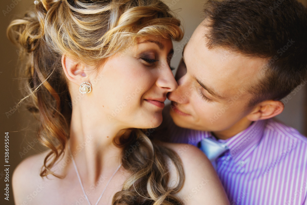 Groom hugs bride from behind and kisses her in rays of morning sun Groom hugs bride from behind and kisses her in rays of morning sun