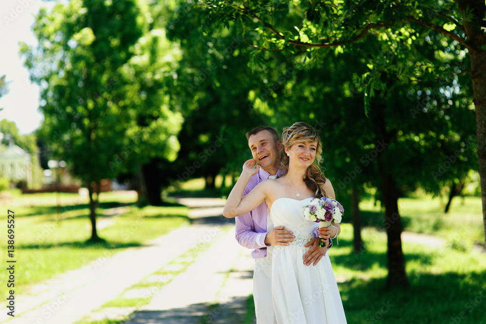 Groom laughs hugging bride from behind on path in sunny park