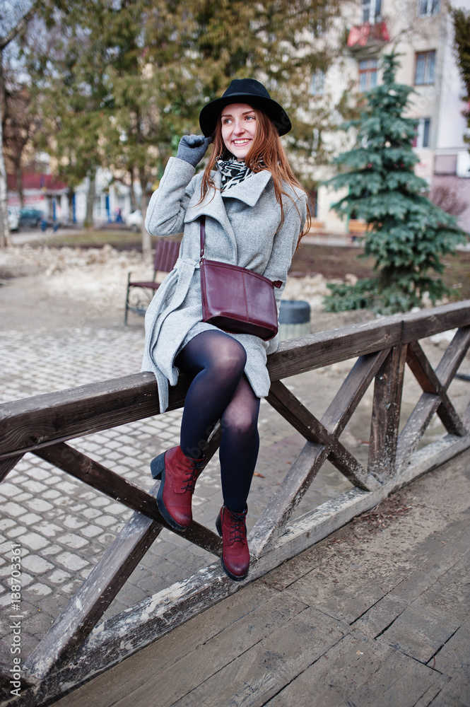 Young model girl in gray coat and black hat with leather handbag on shoulders posed against wooden beams at street of city. Young model girl in gray coat and black hat with leather handbag on shoulders posed against wooden beams at street of city.