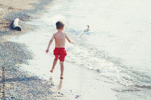 Funny adorable white Caucasian one young little boy in red swim shorts running on beach by water ocean sea with seagull, view from back, emotional lifestyle summer mood