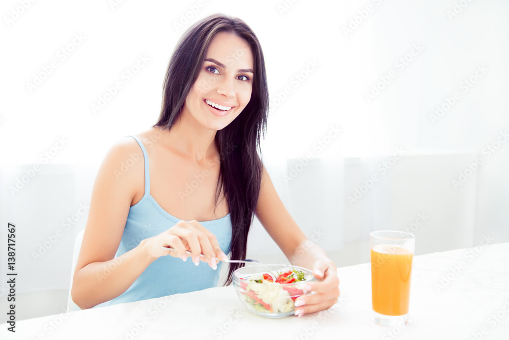 A cheerful young woman eating fresh organic salad and drinking orange juice for breakfast