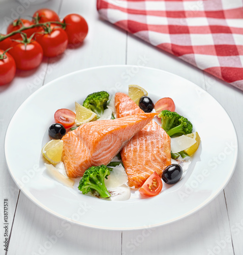 Baked salmon steak grilled with vegetables, olives, lemon and sauce on a white plate on a light wooden background.