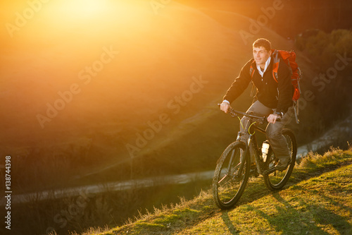 Man with red backpack riding on the mountain bicycle on a trail with bright sun on the sunset photo