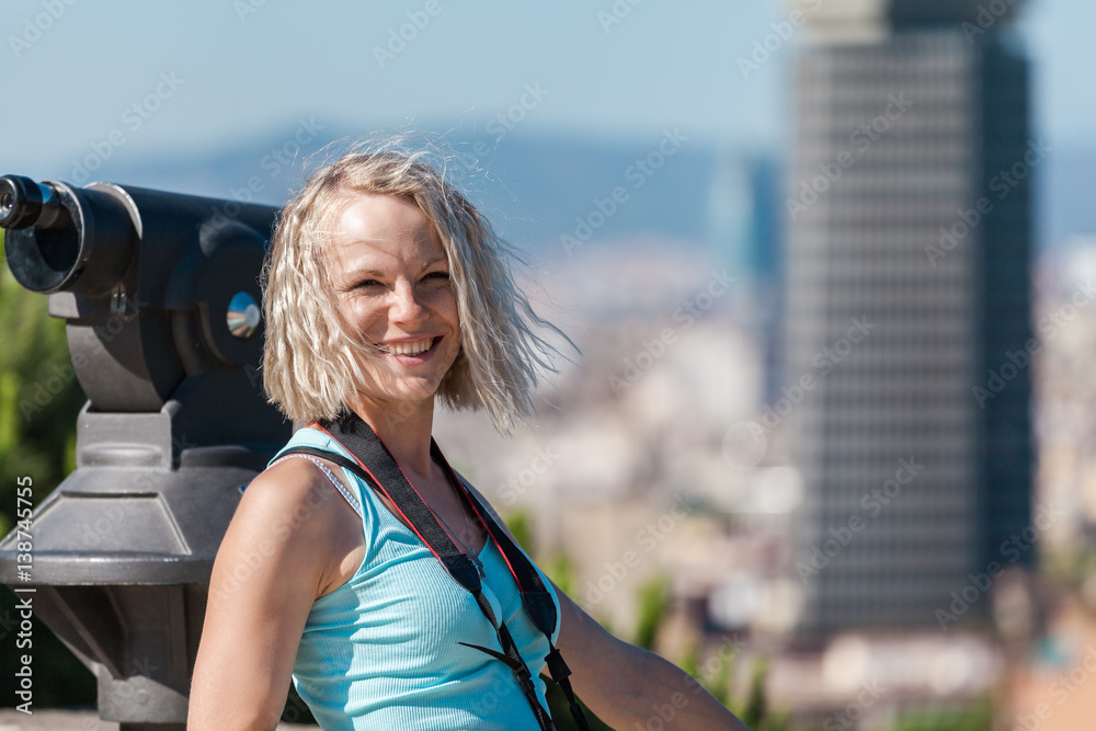 Female traveler standing with camera against the background of the port of Barcelona. Spain. Cute, smiling blond girl with curly hair in a blue t-shirt.